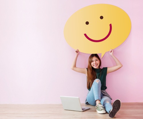 girl holding smiley face while on laptop on pink background